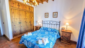 A bedroom with a large wooden wardrobe, a bed with a blue patterned bedspread, two bedside tables with lamps, and two framed pictures on the wall. - Mansion Piedras Blancas Colmenar by Ruralidays (Málaga)