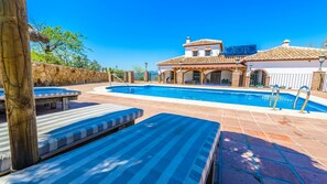 A swimming pool with lounge chairs, a stone wall, and a building with a tiled roof in the background. - Mansion Piedras Blancas Colmenar by Ruralidays (Málaga)