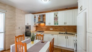 A modern kitchen with white cabinets, a wooden accent wall, a dining table with chairs, and a built-in stove. - Casa Jazmín Azul Cómpeta by Ruralidays (Málaga)