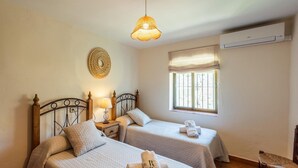 A bedroom with two beds, a bedside table with a lamp, a window with blinds, and a wall-mounted air conditioner. - Casa Jazmín Azul Cómpeta by Ruralidays (Málaga)