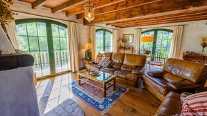 A cozy living room with leather sofas, a glass coffee table, and a wooden floor. Large windows with green frames allow natural light to fill the space. - Hidalgo de Arenas by Ruralidays (Málaga)