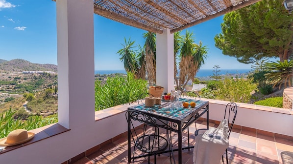 Una terraza con una mesa preparada para dos, con vistas a un paisaje pintoresco con árboles y el mar.