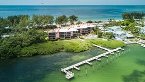 An aerial view of a coastal hotel with a dock extending into the water, surrounded by lush greenery and a clear view of the ocean.