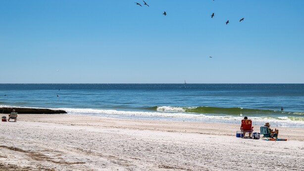 A beach with people relaxing on chairs, a sailboat in the distance, and birds flying overhead.