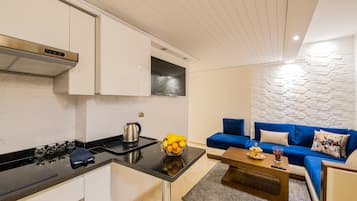 Een moderne keuken met witte kasten, een zwart aanrecht en een ingebouwde kookplaat. Er staat een blauwe bank en een salontafel met een schaal fruit en een afstandsbediening.