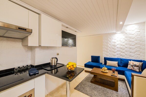 A modern kitchen with white cabinets, a black countertop, and a built-in stove. There is a blue sofa and a coffee table with a bowl of fruit and a remote control. - Marrakech INN (Marrakech)