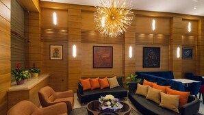 Un área de estar moderna con paneles de madera, un sofá negro con cojines naranja y beige, una mesa de centro redonda con un centro de mesa floral y una lámpara de araña original.