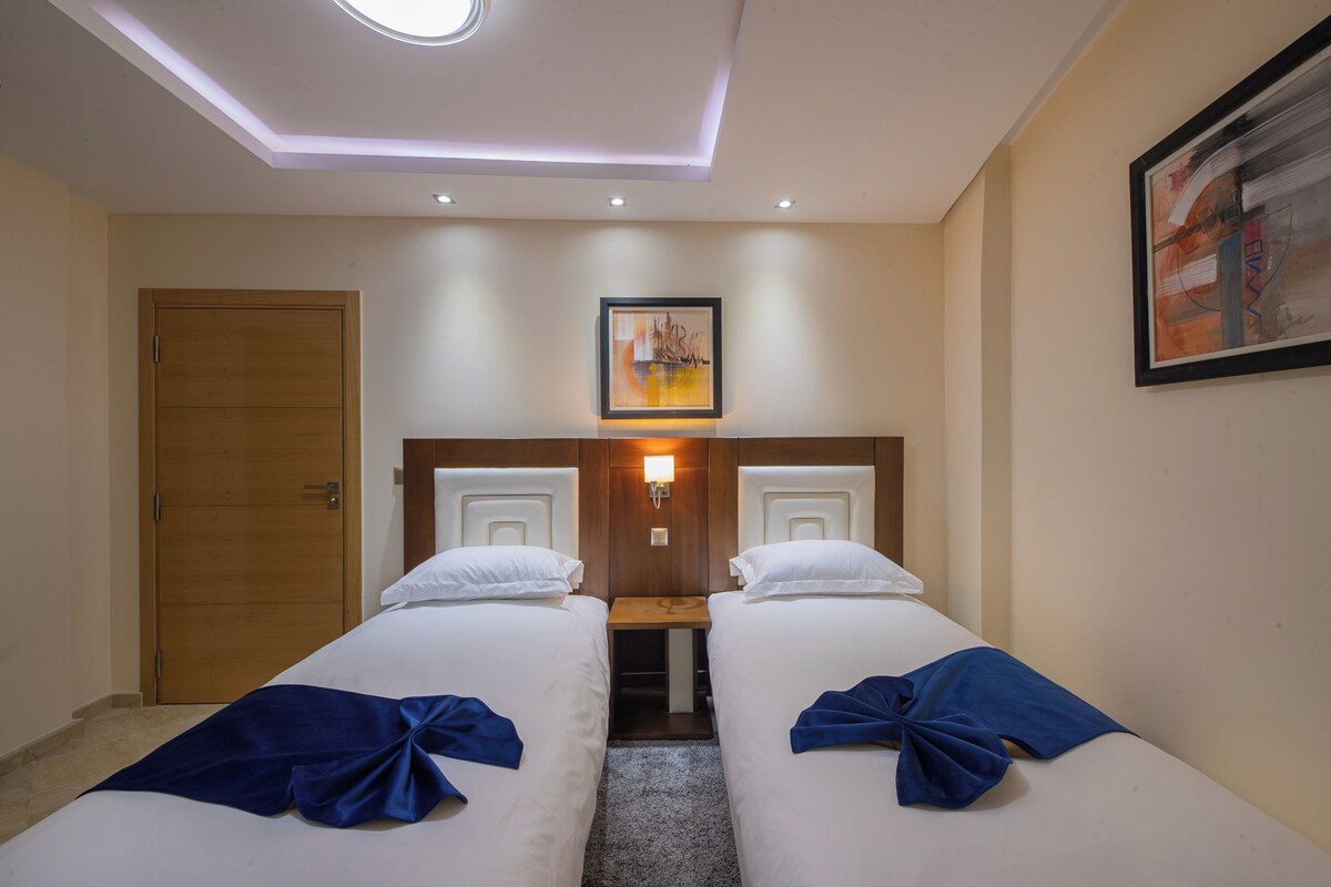 Quarto de hotel com duas camas de solteiro, porta de madeira, luminária no teto e um quadro na parede.