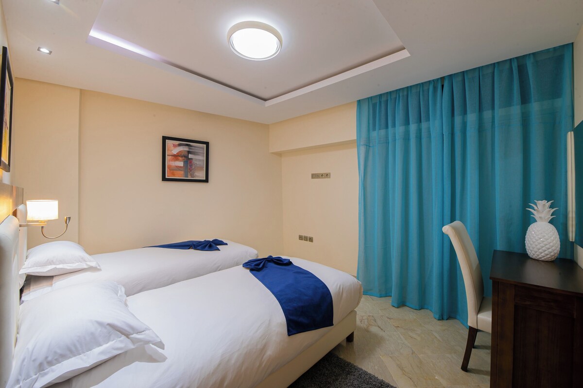 Ett hotellrum med två sängar, ett skrivbord, en stol, en ananasformad dekoration och ett stort fönster med blå gardiner.