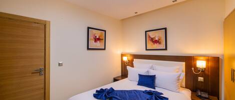Habitación de hotel con una cama grande, una puerta de madera, dos lámparas fijadas a la pared y dos obras de arte enmarcadas.