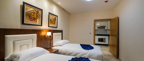 A hotel room with two beds, a wooden headboard, a wall-mounted light, and a door leading to a kitchenette.
