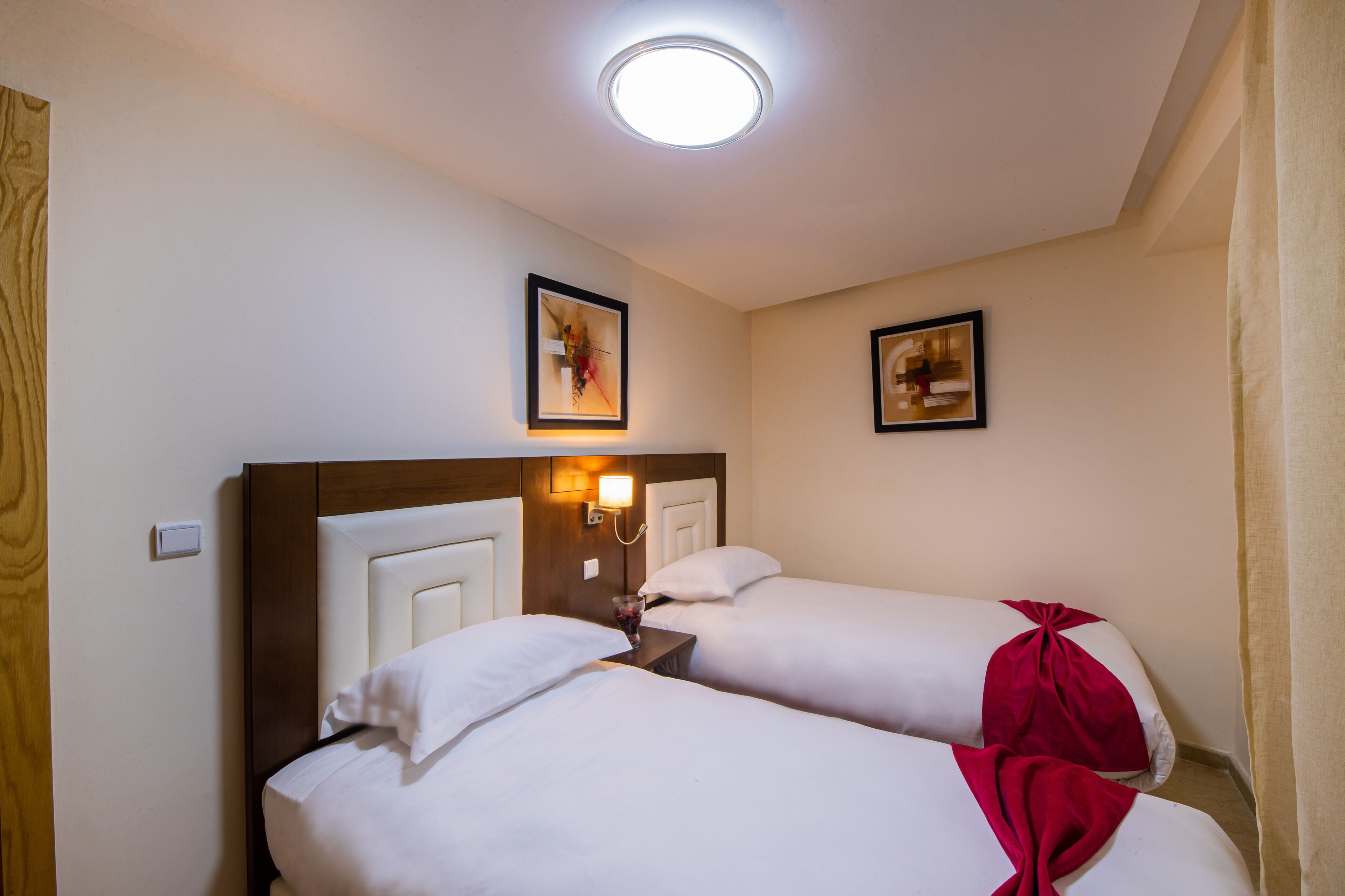 A hotel room with two beds, a nightstand, a wall-mounted lamp, and two framed pictures.