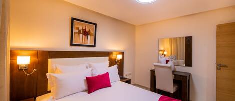 Quarto de hotel com uma cama grande, duas mesas de cabeceira com luminárias, uma escrivaninha com cadeira, um espelho e uma porta.