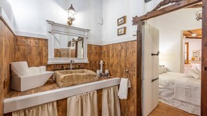 Une salle de bain rustique avec un lavabo en pierre, un lambris en bois, un miroir et un lit recouvert d’une courtepointe blanche.