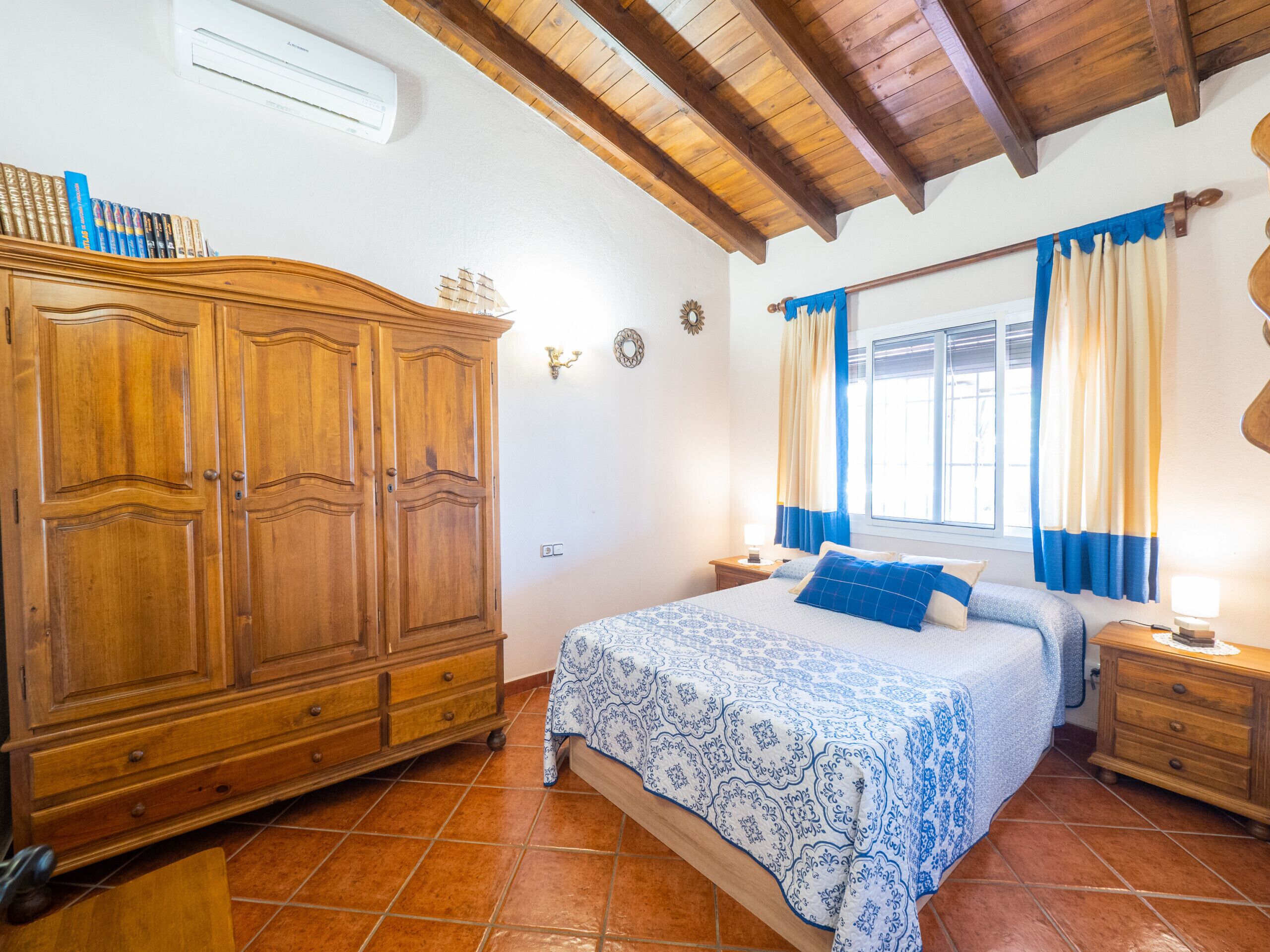 A bedroom with a wooden wardrobe, a bed with a patterned bedspread, a nightstand, and a window with curtains.