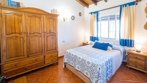 A bedroom with a wooden wardrobe, a bed with a patterned bedspread, a nightstand, and a window with curtains. - Cubo's Finca Los Dragos. Pool, wifi and AC/heating (Alhaurín el Grande)