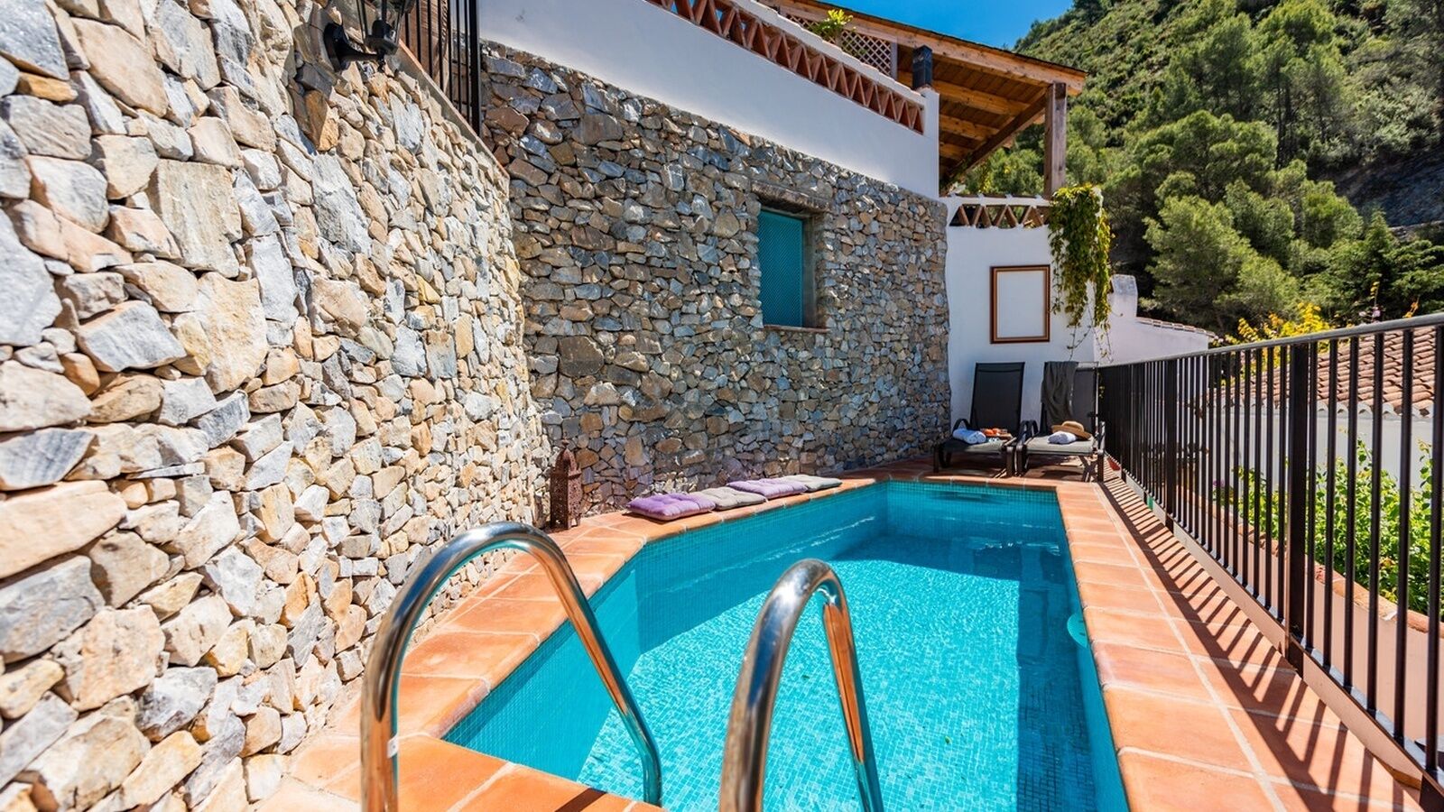 A stone-walled pool with a metal ladder, surrounded by a tiled deck and a metal fence.
