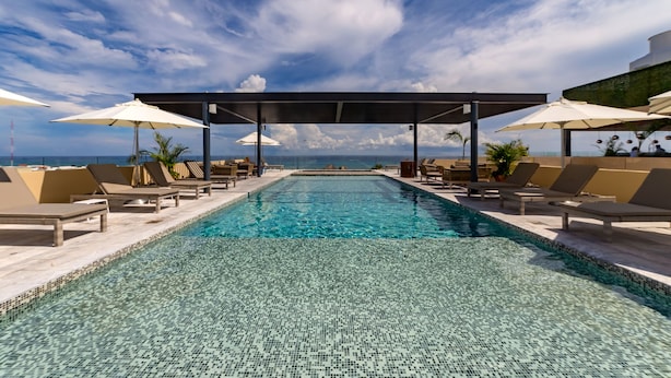 A clear pool with lounge chairs and umbrellas, a covered seating area, and a view of the ocean. - Singular Joy Vacation Rentals (Playa del Carmen)