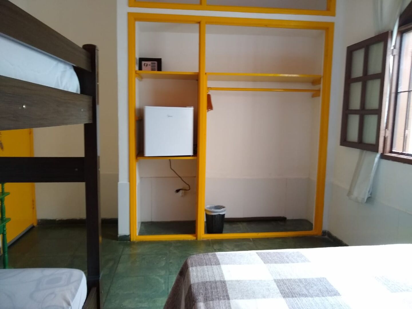 Una habitación con literas, un armario con un pequeño electrodoméstico blanco, un cubo de basura y una ventana con cortinas.