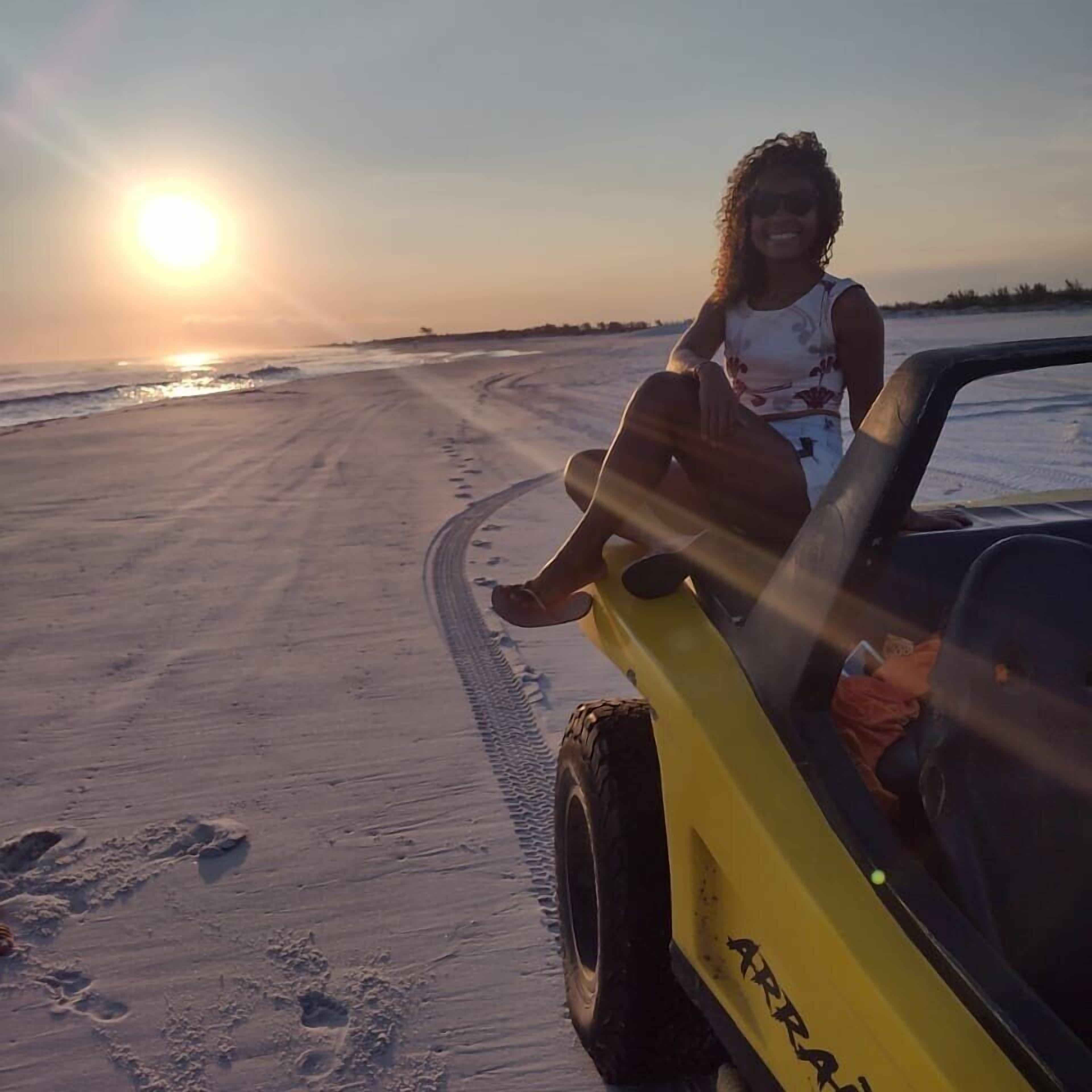 Une personne assise sur un véhicule jaune, sur une plage de sable, avec des empreintes de pas qui mènent à l’eau, au coucher du soleil.