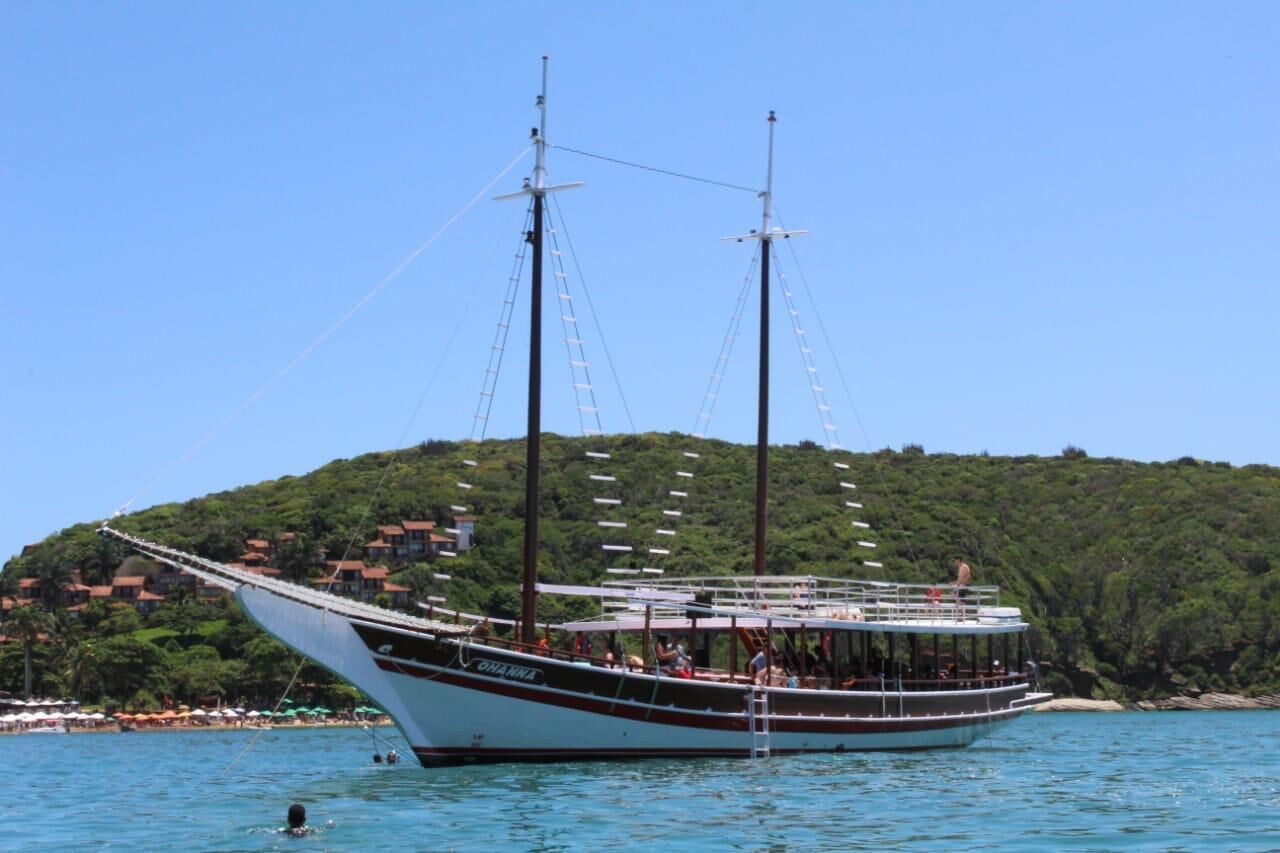 Un barco grande con varias cubiertas y una estructura de cabina, amarrado cerca de una playa con una colina al fondo.