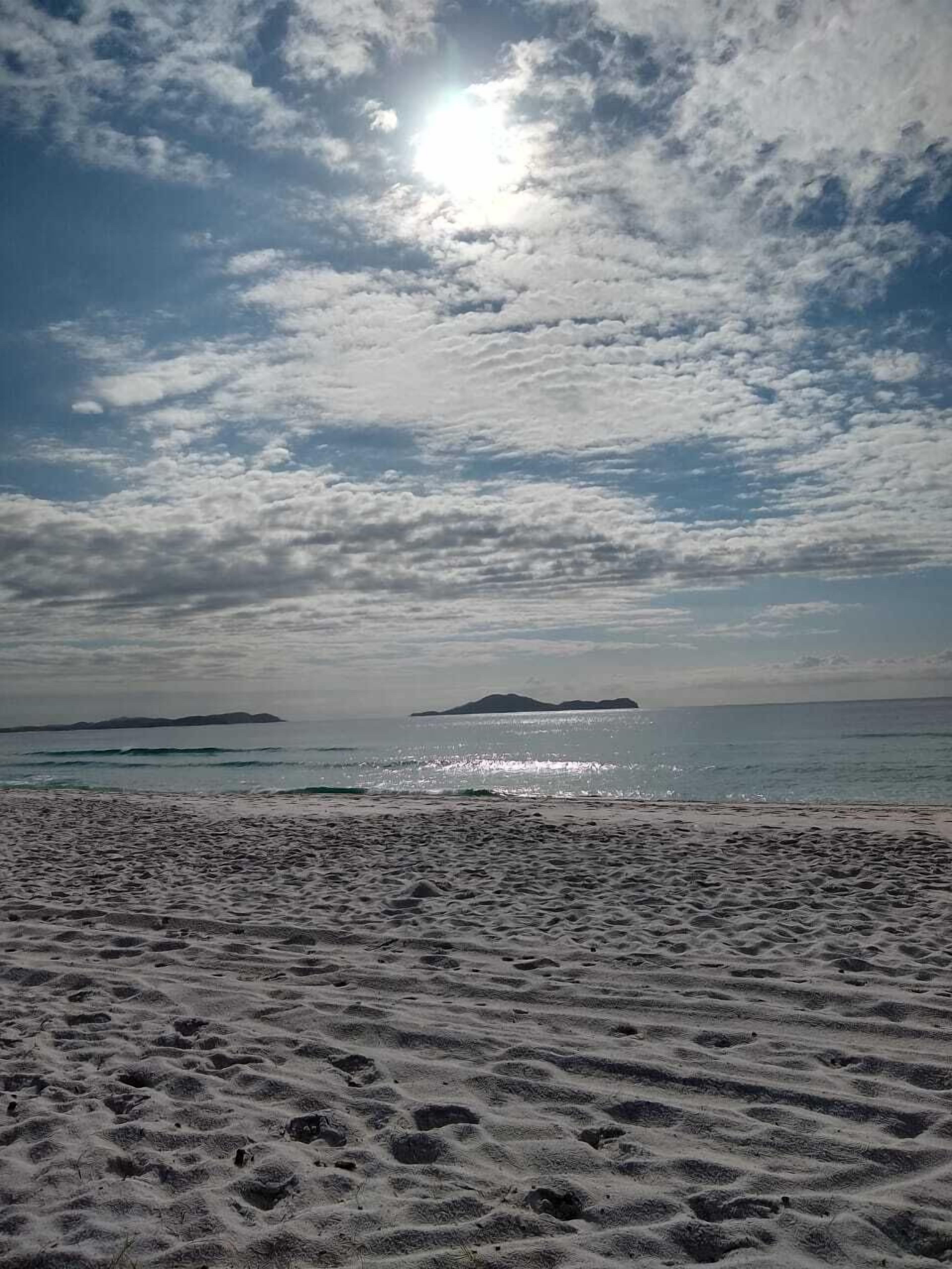 Platja amb empremtes a la sorra, mar tranquil·la i una illa llunyana sota un cel amb núvols dispersos.