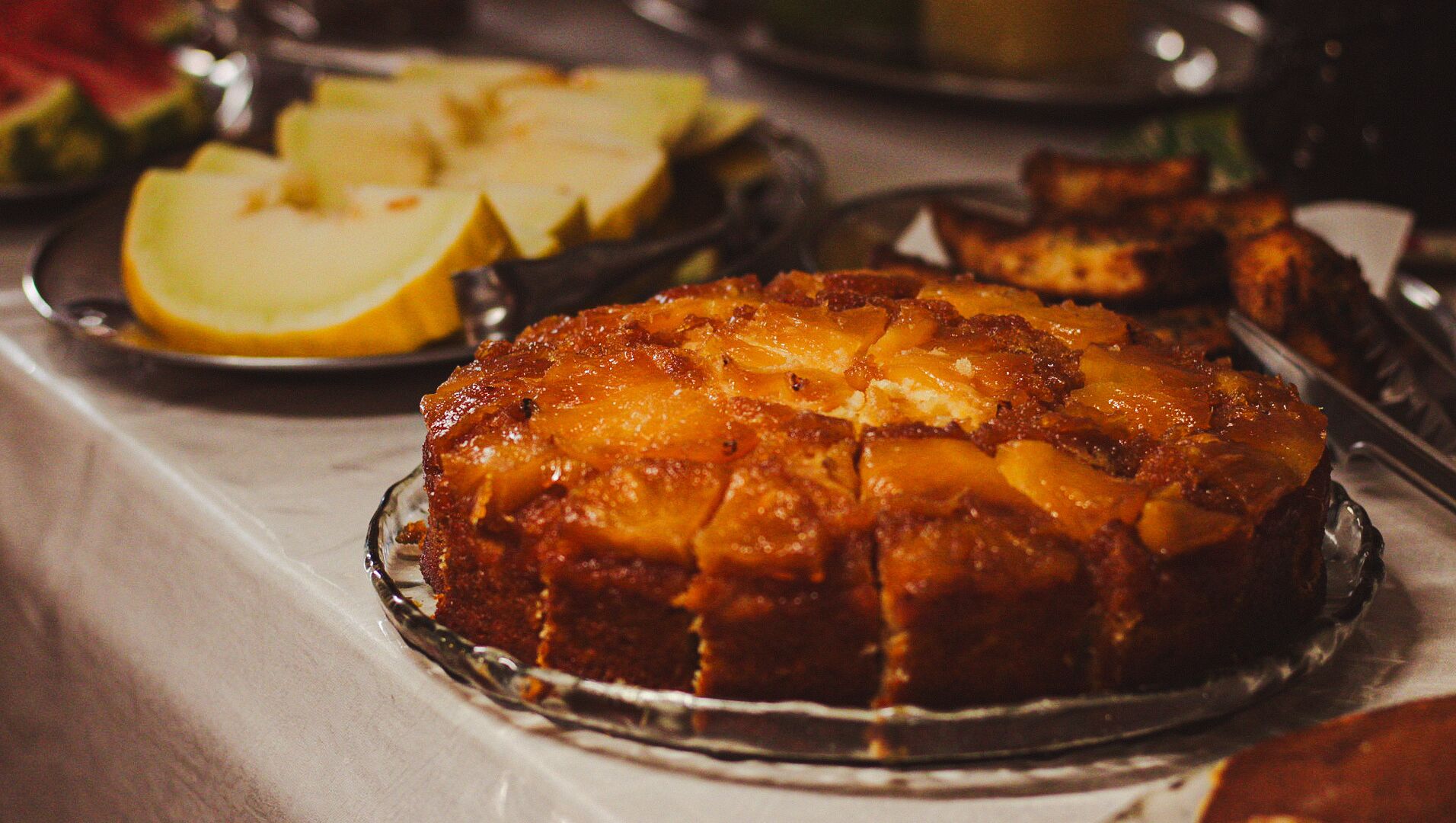 A dessert dish with a caramelized top, accompanied by sliced apples and a glass of beverage on a table.