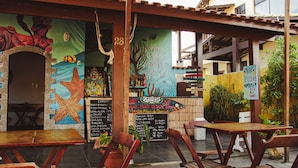 An outdoor dining area with wooden tables and chairs, a mural with marine life, and a chalkboard menu listing food and drink prices. - Beach Hostel & Suites, Cabo Frio (Cabo Frio)