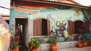 A building with a mural of an underwater scene, including a submarine and marine life. There are three windows with shutters, and potted plants are placed in front of the windows. - Beach Hostel & Suites, Cabo Frio (Cabo Frio)