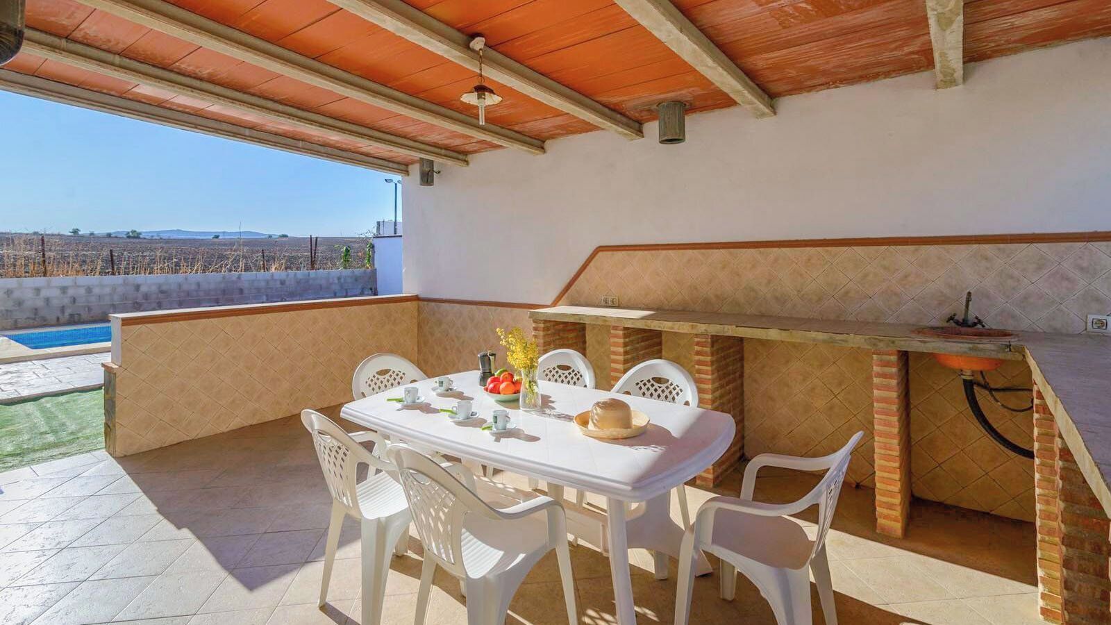 Un patio al aire libre con una mesa lista para comer, rodeado de sillas y una barra de ladrillo con un lavamanos.