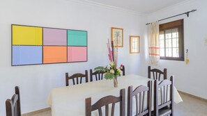 Ein Essbereich mit einem Tisch, der für sechs Personen gedeckt ist, umgeben von Holzstühlen. Ein farbenfrohes abstraktes Gemälde hängt an der Wand, und ein Fenster mit Vorhängen ist sichtbar.