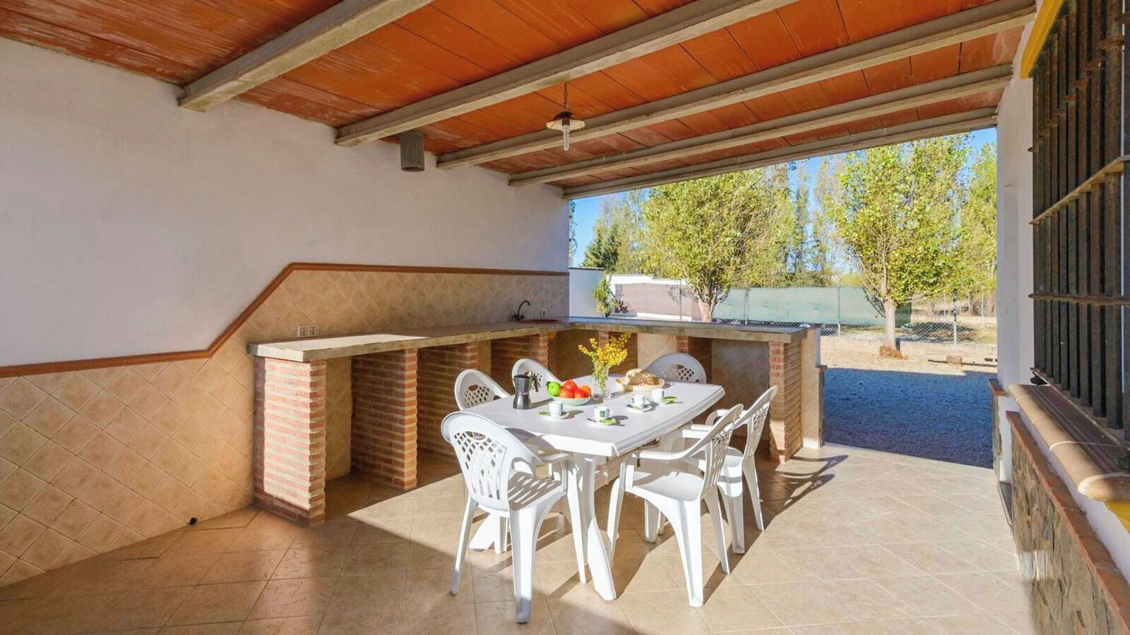 Un patio al aire libre con una zona de parrillada de ladrillo, sillas de plástico blancas y una mesa lista para comer.
