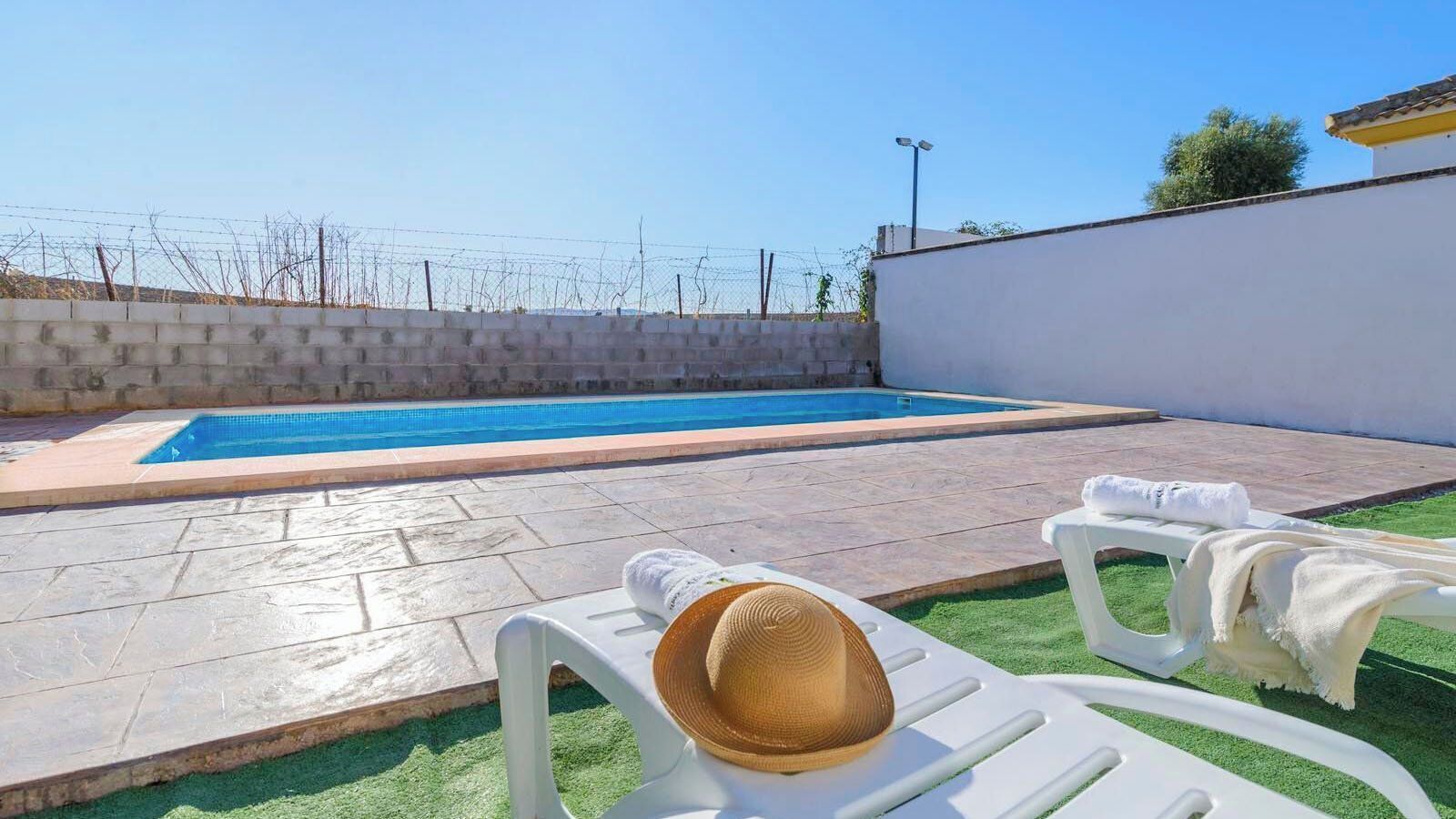 Une piscine avec une terrasse en pierre, deux chaises longues blanches et un mur blanc.