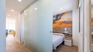A modern hotel room with a wooden floor, a bed with a white bedspread, a nightstand, and a wall mural of a sunset over the ocean.