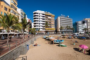 Un hotel frente a la playa con varios pisos, terraza exterior y barcos en la arena.