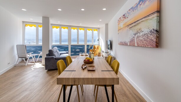 A modern dining area with a wooden table, yellow chairs, and a view of the ocean through large windows.
