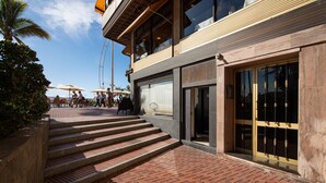 A hotel entrance with a brick pathway leading up to the doors, a palm tree, and outdoor seating with umbrellas.