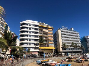 Un hotel con varios pisos, balcones y una zona de playa con barcos y sombrillas.