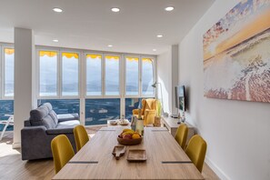 Una sala de estar moderna con una mesa de comedor, sillas amarillas, un sofá gris y un ventanal con vistas a la ciudad.