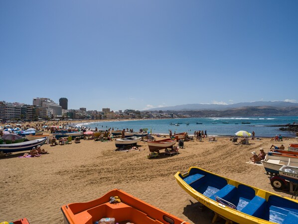 Una playa con barcos y sombrillas, cielo despejado y edificios al fondo.