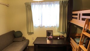 Um quarto pequeno e acolhedor com piso de madeira, um sofá, uma cama beliche, uma mesa com porta-retrato e uma pequena luminária, além de uma janela com cortinas.