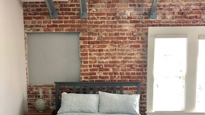 Une chambre avec un mur en briques, un lit avec une literie blanche, une table de chevet avec une lampe et une fenêtre avec des stores.