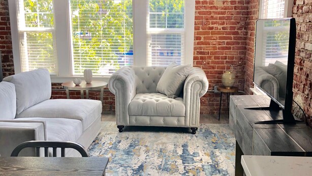 Un salon avec un mur en briques, un canapé blanc, un fauteuil à capitons, une table basse, un téléviseur et un tapis à motifs.