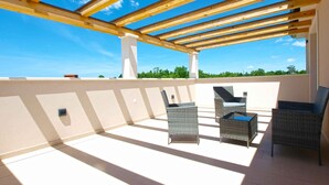 Un espacio al aire libre techado, con vigas de madera, suelo de baldosas y mobiliario de mimbre.