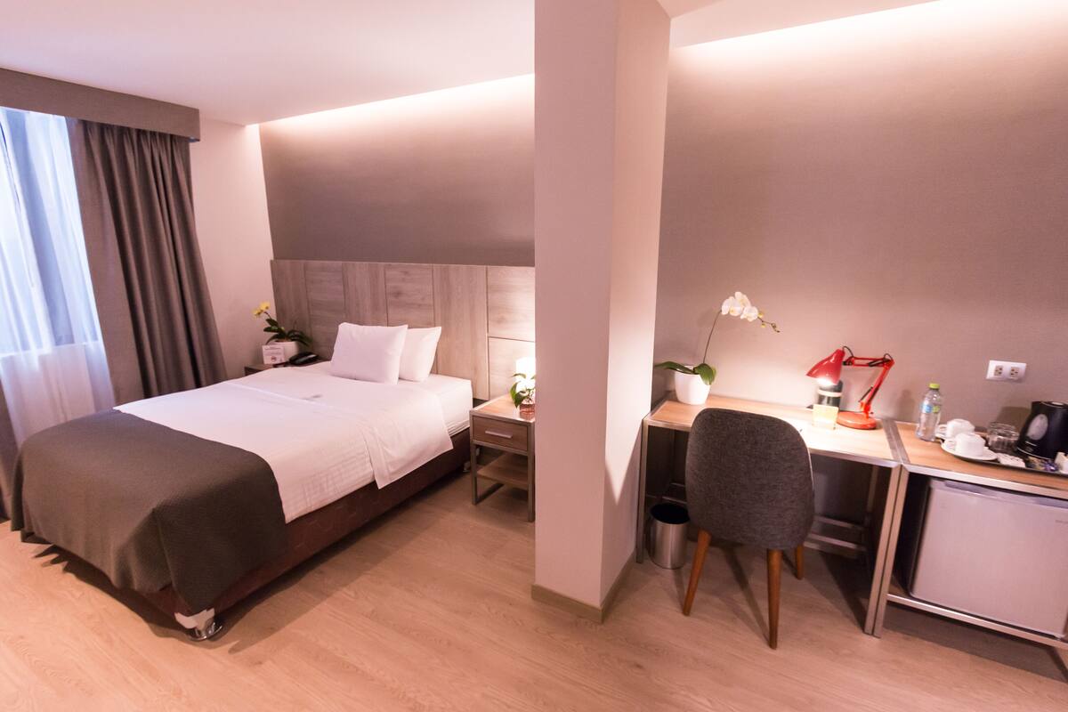 A modern hotel room with a bed, bedside table, lamp, desk, chair, and a small kitchenette.