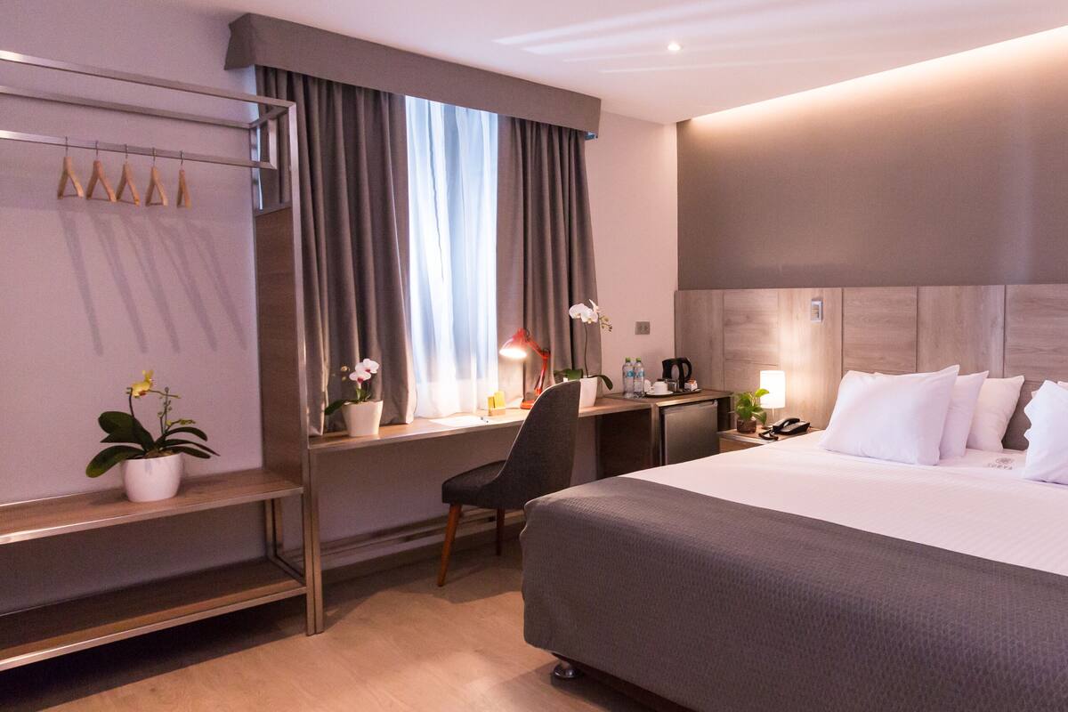 A hotel room with a large bed, a desk with a chair, a nightstand, and a potted plant.