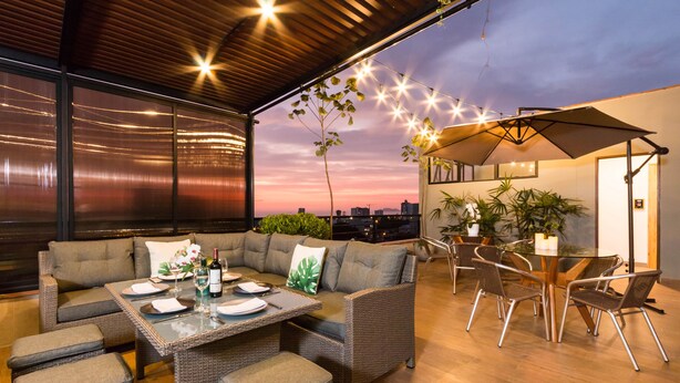 An outdoor seating area with a sofa, table, and chairs, a balcony with a view of the city skyline, and string lights. - Surya Lima (Lima)