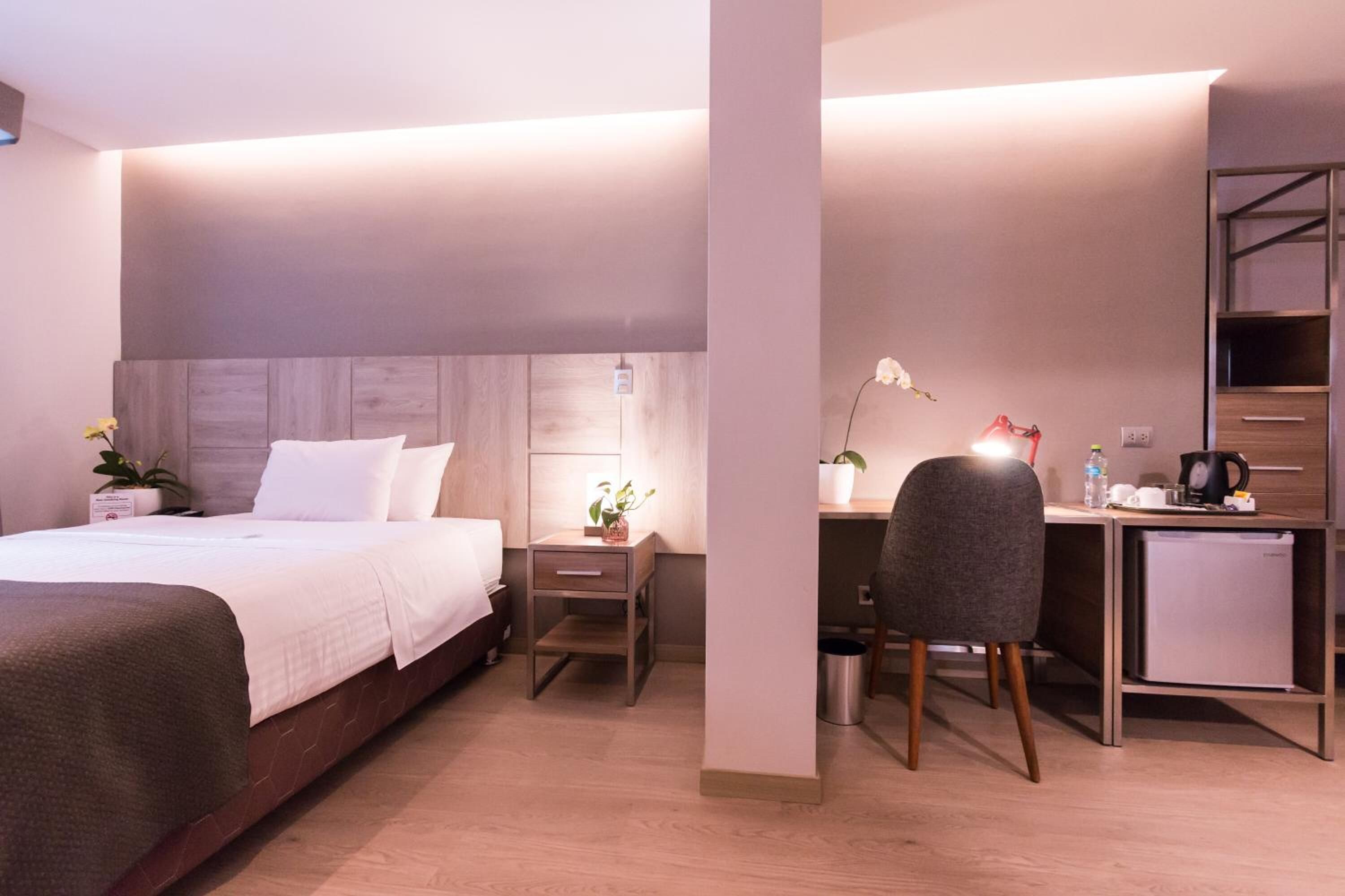 A modern hotel room with a bed, bedside table, lamp, chair, desk, and a small refrigerator.