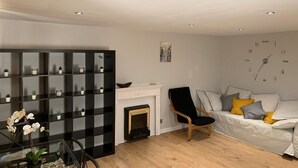 A living room with a fireplace, a black shelving unit, a white sofa with cushions, and a clock on the wall. - Luks Apartments (London)