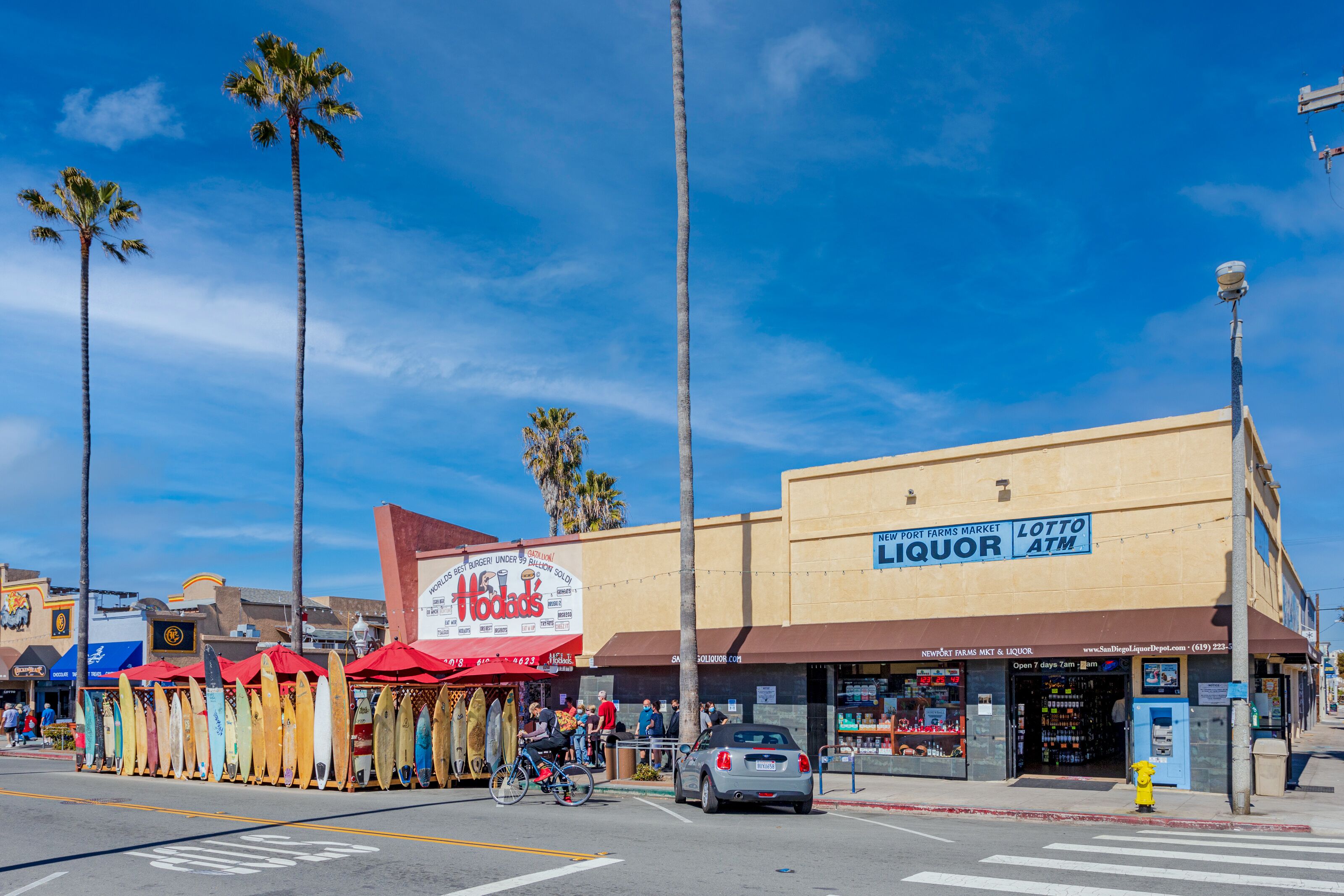 Une vue de rue avec une boutique de vente de boissons alcoolisées, un magasin de location de planches de surf et des palmiers.